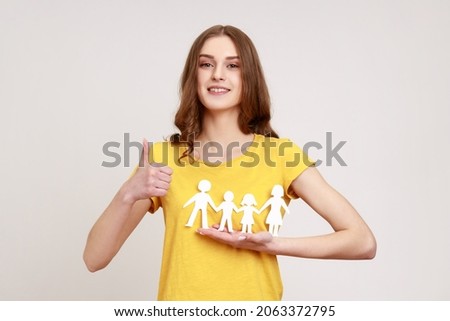 Portrait of happy satisfied female of young age holding paper family chain and showing thumb up, looking at camera with toothy smile. Indoor studio shot isolated on gray background.