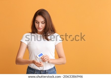 Young woman with infrared thermometer measuring her temperature on color background