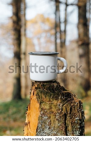 Camping metal white mug mock up standing on tree stump in woods outdoors. Enamelled balnk cup with empty space for branding or logo. Vertical photo.