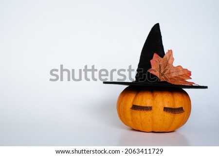 Creative small orange pumpkin with black witch hat eyelashes and autumn leaf on white isolated background. Halloween and Thanksgiving season concept