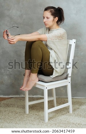 depressed view of a girl taking off her glasses, sitting on a white chair against the background of an old cracked wall