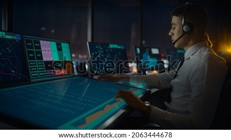 Male Air Traffic Controller with Headset Talk on a Call in Airport Tower. Office Room is Full of Desktop Computer Displays with Navigation Screens, Airplane Departure and Arrival Data for the Team. Royalty-Free Stock Photo #2063444678