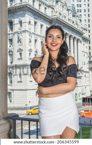 Portrait of College Student. Dressing in black, short sleeve top, white short wrap skirt, a young pretty lady with long curly hair is standing in the front of office building, relaxing, smiling. 