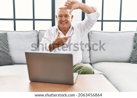 Senior man using laptop at home sitting on the sofa smiling making frame with hands and fingers with happy face. creativity and photography concept. 