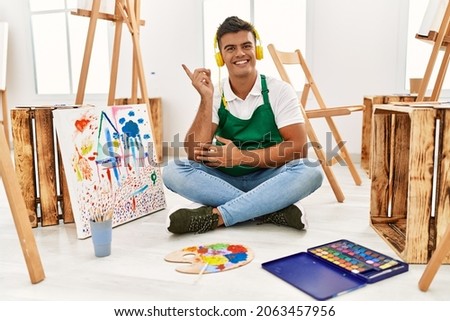 Young hispanic man at art studio with a big smile on face, pointing with hand and finger to the side looking at the camera. 