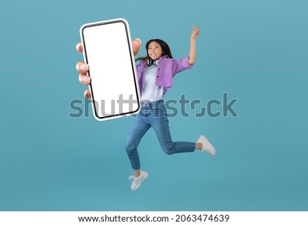 Excited asian lady showing empty smartphone screen while jumping up over blue studio background, collage, full body length, banner. Happy woman recommending cool mobile app or website