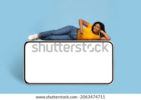 Pretty young african american woman in stylish casual outfit chilling on huge smartphone with empty screen, pointing down and smiling at camera, entertaining mobile app concept, mockup