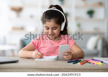 Online Lesson. Cute Little Girl In Headphones Using Smartphone For Study At Home, Adorable Arab Female Child Sitting At Table In Room, Holding Cellphone And Writing In Notepad, Closeup Shot
