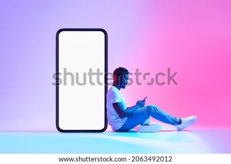 Young black man sitting next to huge smartphone with empty screen, advertising new mobile application or website, using modern gadget in neon light. Cellphone display mockup Royalty-Free Stock Photo #2063492012