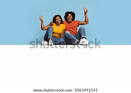Carefree millennials black man and woman sitting on empty white board for your text or advertisement together, embracing, showing peace gesture and smiling at camera, blue background, copy space