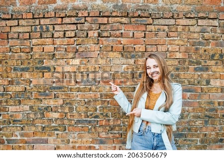 Happy young blonde woman pointing sideways, portrait. Brick wall background. Banner. Photo for advertising and layout. 