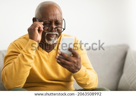 Poor Eyesight. Senior Black Man Using Smartphone Wearing Eyeglasses Sitting On Sofa At Home. Bad Vision, Eyes Health And Healthcare Problems Concept. Selective Focus Royalty-Free Stock Photo #2063508992