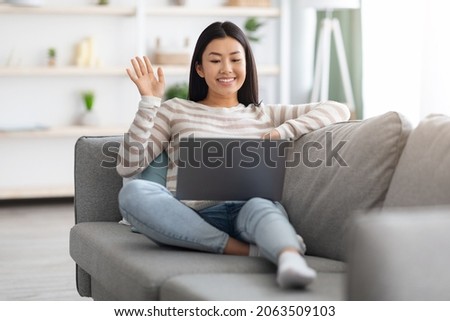 Distant Communication. Beautiful Asian Lady With Laptop At Home Making Video Call To Friends Or Family, Cheerful Young Korean Female Sitting On Couch And Waving Hand At Web Camera, Copy Space