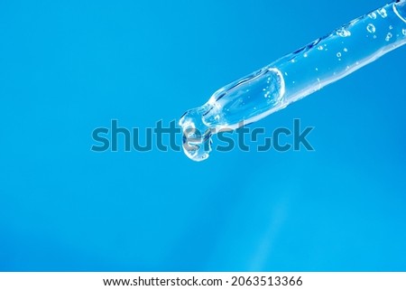 Glass pipette with facial serum, bubbles inside and falling drop on light blue background macro. Modern cosmetic product advertising. Medical research, innovation concept. Dropper with gel closeup.