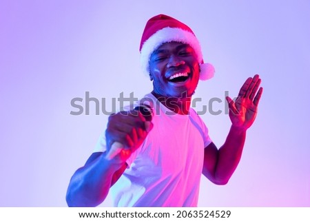 Christmas music, New Year concert. Young black man in Santa hat singing karaoke into microphone, dancing and having fun on Xmas party in neon light. Millennial guy performing song on holiday night