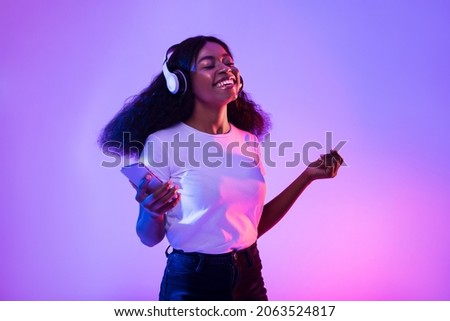 Happy African American woman with headphones and smartphone listening to music and dancing in neon light. Young black woman moving to favorite song, enjoying cool playlist Royalty-Free Stock Photo #2063524817