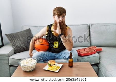 Caucasian man with long beard holding basketball ball cheering tv game hand on mouth telling secret rumor, whispering malicious talk conversation 