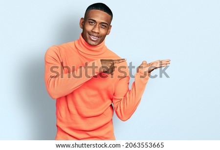 Young black man wearing orange turtleneck sweater amazed and smiling to the camera while presenting with hand and pointing with finger. 