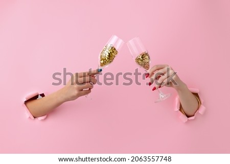 Cheers. Two female hands holding champagne, wine glass with confetti breaks through pink paper background. Copy space for ad, design, text. Concept of holidays, celebrations and greeting. Faceless.