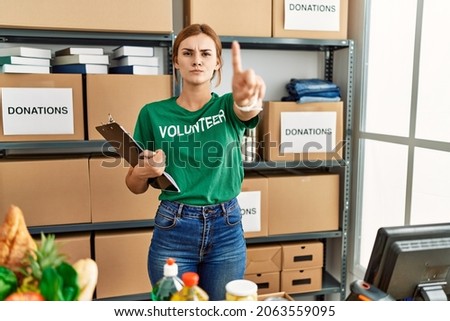Young brunette woman wearing volunteer t shirt at donations stand pointing with finger up and angry expression, showing no gesture 