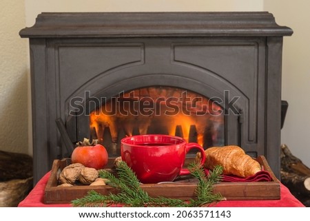 Christmas and winter holidays home interior; Plate with two glasses of red wine, fruits, nuts, fir twigs, two wicker chairs in front of burning iron stove