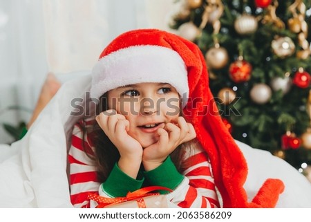 little girl in a New Year's red hat with a Christmas gift. She lies on the bed, hugging a box against the background of a Christmas tree.