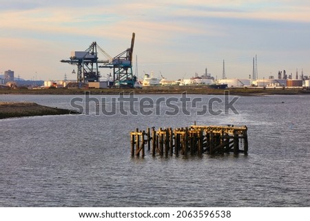 An Old wooden Jetty in the River Tees with Cranes and a Oil Refinery in the background, Middlesbrough, North Yorkshire, England, UK. Royalty-Free Stock Photo #2063596538