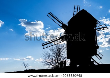 silhouette of an old wooden mill against the background of a clear blue sky with a couple of clouds, several contours of people 