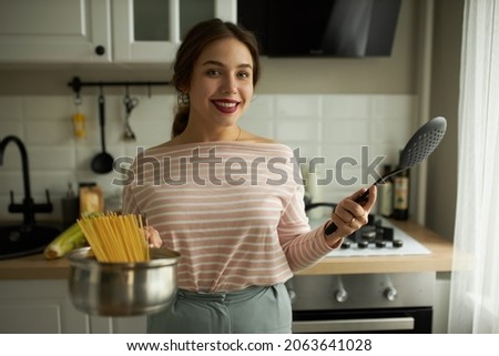 Indoor picture of young beautiful elegant woman wearing red lipstick, holding saucepan with spaghetti and black spotted spoon posing against modern cozy kitchen in stylish casual clothes