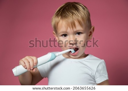Smiling caucasian little boy cleaning his teeth with electric sonic toothbrush on pink background.