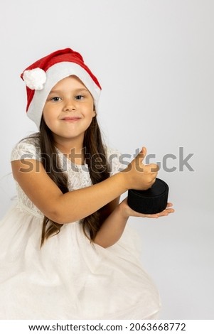 portrait of laughing, joyful girl in Santa Claus hat holding smart music column or station in hands and giving a thumbs up, isolated on a white background, the concept of a high-tech New Year gift. 
