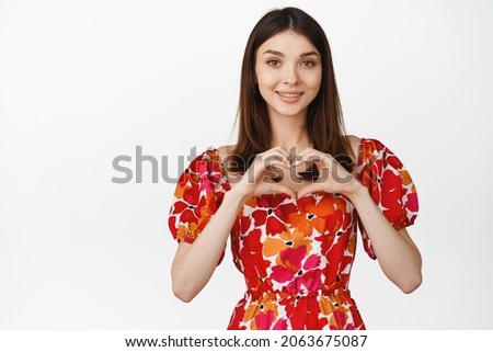 Cute and feminine girl shows heart gesture, smiling and looking with tender face expression at camera, white background