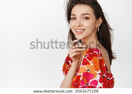 Beauty. Young hispanic woman with hair flying in air, gazing at camera and touching fresh, clean skin with light makeup on, standing over white background