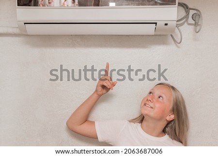 Cute teenage girl blonde European appearance points a hand finger at the air conditioner on the wall in the room.
