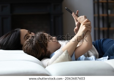 Cute boy and young mom using smartphone, resting on couch together, taking home selfie, texting, chatting online, looking at screen. Happy mother and kid holding mobile phone, relaxing on sofa