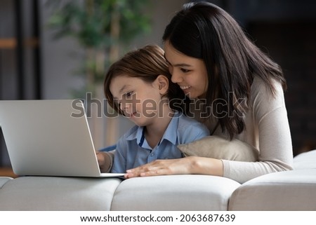 Happy millennial Asian mom and gen Z son boy watching movie, media content on laptop computer, using app, service, chatting online. Mother and son shopping, booking on internet at home