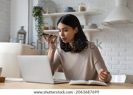 Serious young Asian student woman recording, sending audio message on smartphone, using voice recognition app. Remote employee working at laptop from home giving command to virtual assistant