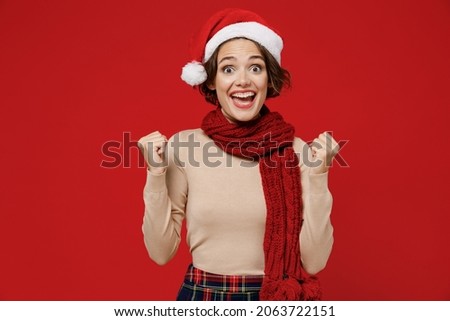 Young oerjoyed excited caucasian woman 20s wear Santa Claus Christmas red hat do winner gesture clench fist isolated on plain red background studio portrait. Happy New Year 2022 celebration concept