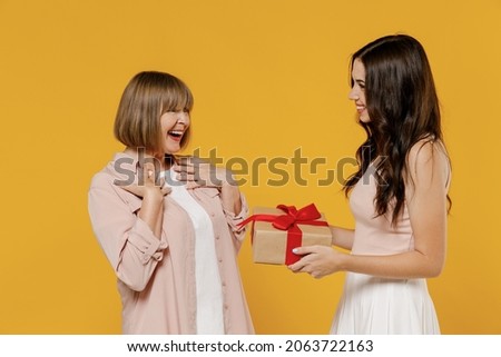 Side view two fun young smiling happy daughter mother together couple women in casual beige clothes gifting birthday present with red ribbon isolated on plain yellow color background studio portrait. Royalty-Free Stock Photo #2063722163