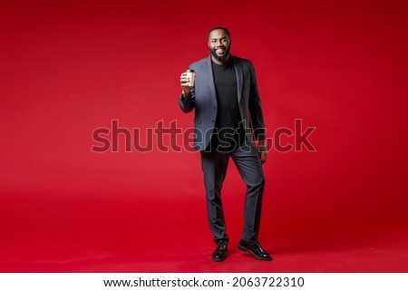 Full length of smiling handsome young black business man 20s in classic jacket suit hold paper cup of coffee or tea looking camera isolated on bright dark red plain color background studio portrait