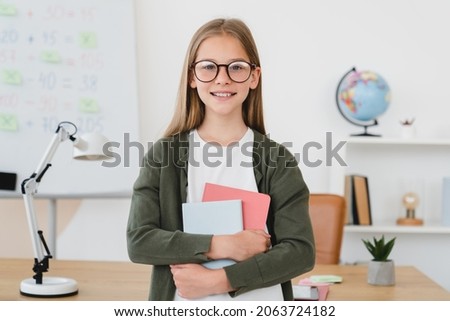 Confident smiling schoolgirl pupil student holding copybooks, standing at school, learning from home remotely, preparing homework online. Royalty-Free Stock Photo #2063724182