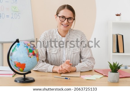Young female maths geography teacher headmaster tutor student lector working at the desk looking at the camera in school classroom. Royalty-Free Stock Photo #2063725133