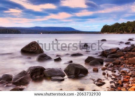 Scenic sunrise over blurred waters of Lake St Clair with wet shore boulders and distant ferry wharf from Platypus bay.