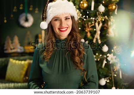 Christmas time. Portrait of happy modern 40 years old woman with Santa hat in green dress near Christmas tree in the modern house. Royalty-Free Stock Photo #2063739107