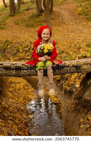 a beautiful happy little blonde girl in a red coat and a red beret is sitting on a bridge across a river in the city's autumn yellow park holding a bouquet of autumn flowers in her hands