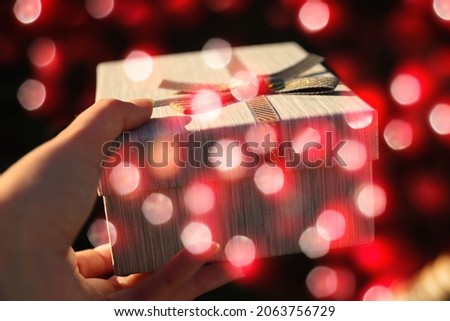 Female hand holding and giving wrapped vintage gift box with ribbon bow on shiny dark red bokeh background. Present box, creative picture. Holiday concept. Glitter backdrop.