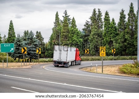 Industrial bonnet bright red classic big rig semi truck transporting commercial cargo in dry van semi trailer turning on the round highway exit with green trees on the side and road signs