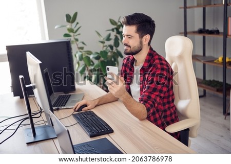 Photo of happy cheerful young man sit table look screen computer hold phone editor indoors inside office