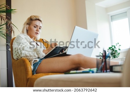 Bottom view of a caucasian woman listening to online lessons. Standing comfortably on the couch with her legs outstretched on the table, noting something in her