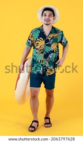 Young Asian man in colorful printed Hawaiian shirt smiles with happiness while holding swim ring. Full body studio portrait on yellow background. Summer holiday travel concept